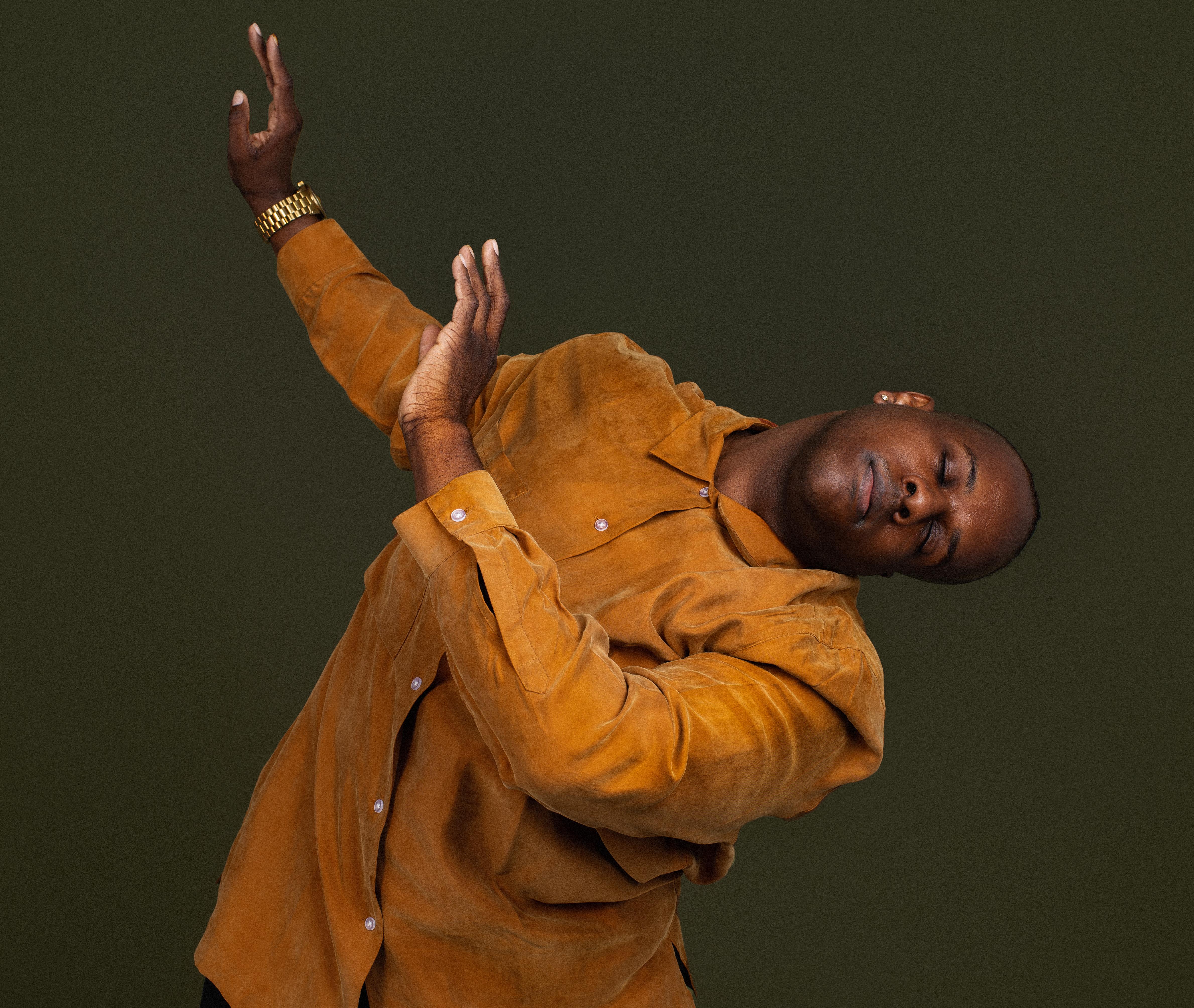 A Black choreographer and dancer is wearing a burnt orange tunic, leaning to the left with his body and gesturing to the right of the frame of the picture